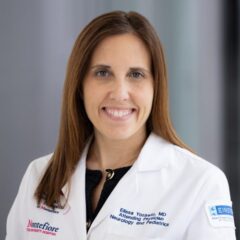 Elissa Yozawitz, MD, in a white medical coat with printed text, stands indoors, smiling warmly at the camera.