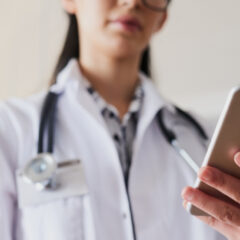 A person in a white coat with a stethoscope around their neck is holding a smartphone, possibly checking the latest updates on the 2024 presidential election.