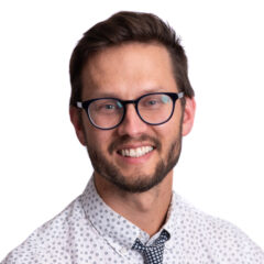 Una persona con cabello corto y castaño, con gafas y una camisa estampada con corbata, sonríe a la cámara sobre un fondo blanco.