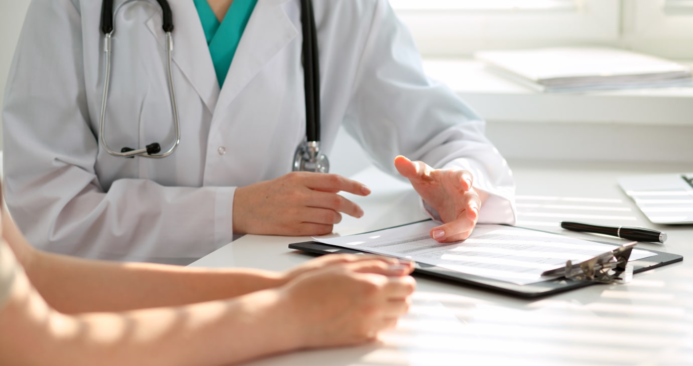 A doctor in a white coat discusses medical documents with a patient at a table in a brightly lit room.