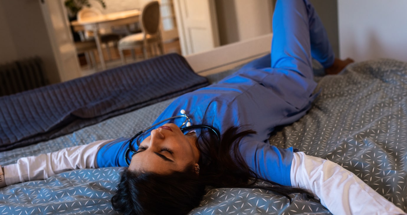 A doctor in blue scrubs lies relaxed on a bed, arms outstretched, stethoscope around neck, finding a moment of peace amidst the mental health crisis.