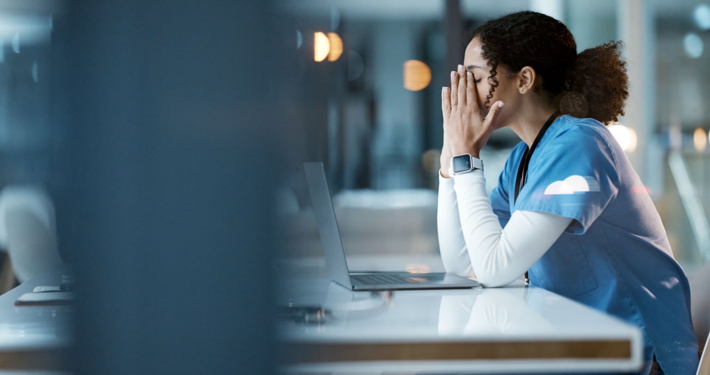 A doctor in scrubs sits at a desk with a laptop, resting their head in their hands, appearing tired and stressed, highlighting the escalating mental health crisis among medical professionals.