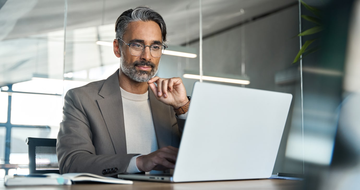 A man with glasses and a beard is sitting at a desk, using a laptop. Wearing a beige blazer, he gazes thoughtfully at the screen, perhaps researching the latest insights into mental health.