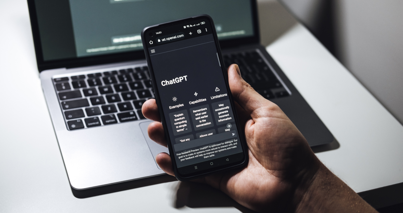 A person holding a smartphone displays the ChatGPT website, hinting at advances in AI communication, while a laptop sits in the background.