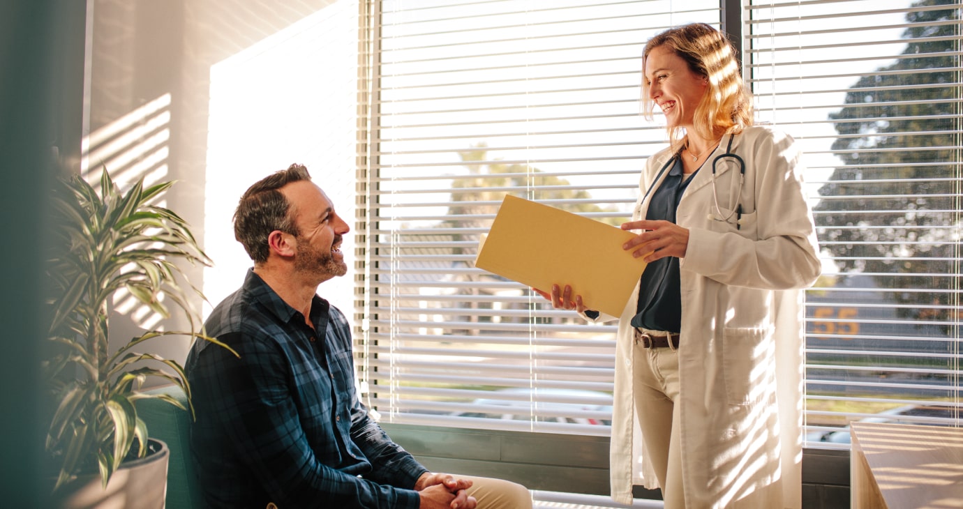 A doctor stands holding a file, discussing patient care with a seated individual in a sunlit office adorned with blinds and a plant.