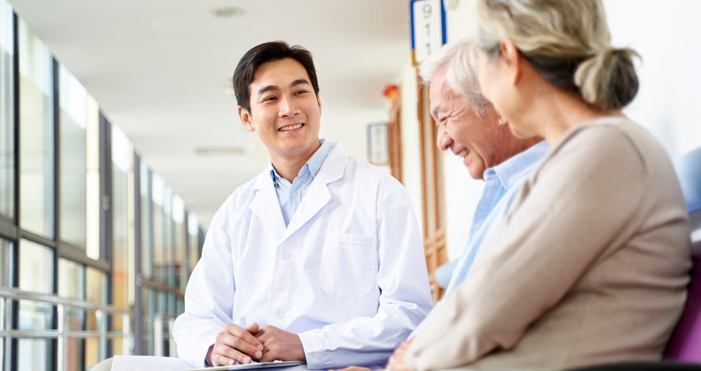 A doctor in a white coat discusses 2025 solutions to improve patient care with two older adults, a man and woman, in a bright hospital corridor.