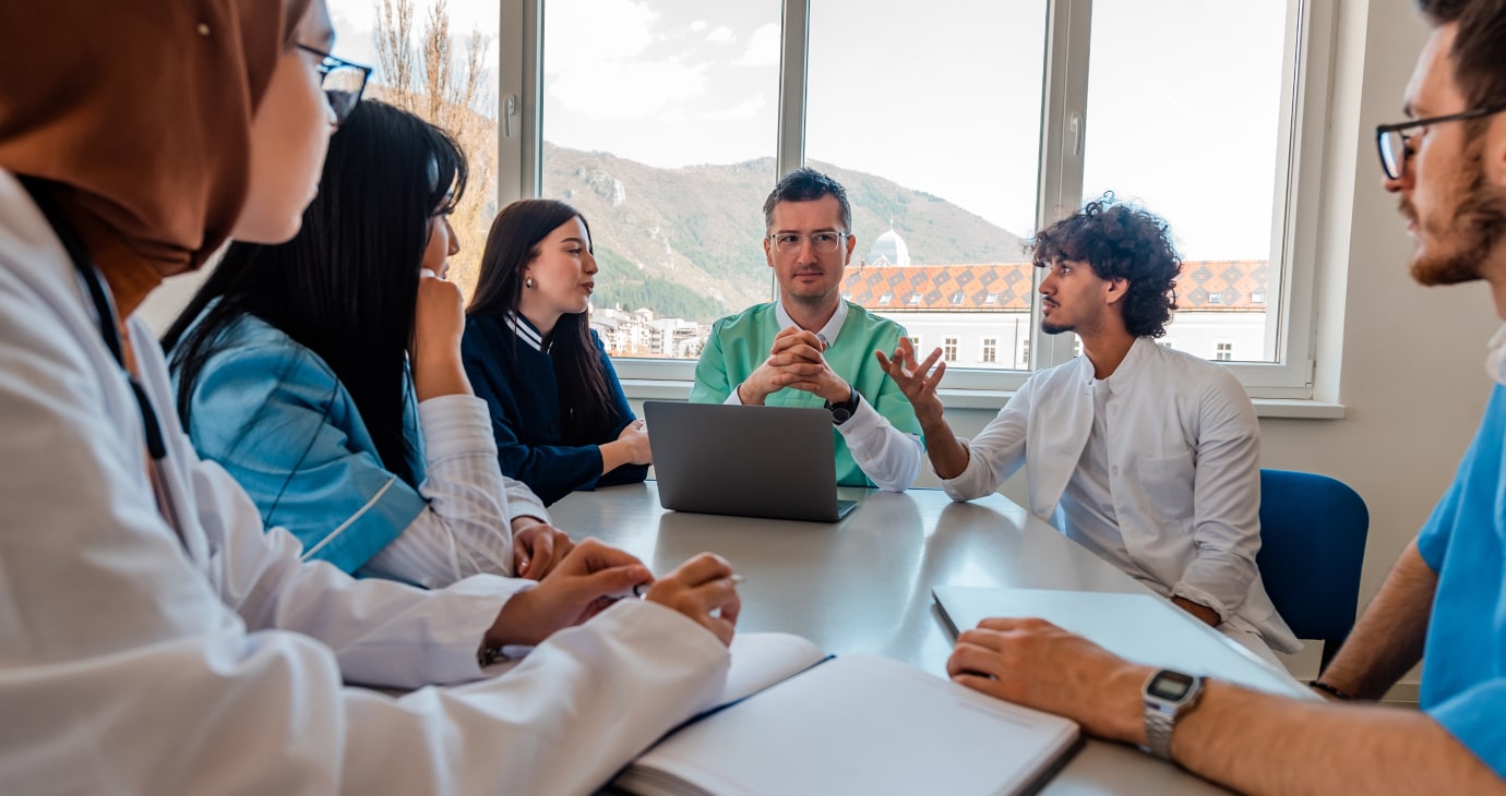 Six healthcare professionals are in a meeting room, seated around a table with a laptop, engaged in a discussion about improving patient care. A window offers a picturesque view of mountains in the background, inspiring their dedication to better outcomes in 2025.