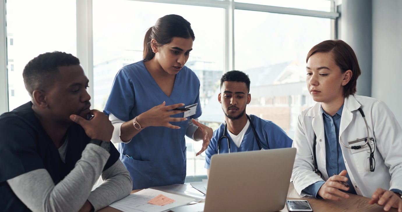 Four healthcare professionals in blue uniforms and white coats engage in collaborative education as they discuss patient records around a laptop in a bright room.