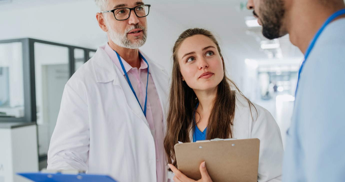 Three healthcare professionals in white coats discuss a matter in a hospital corridor, highlighting collaborative education. They hold clipboards and wear ID badges, embodying modern models of teamwork in medicine.