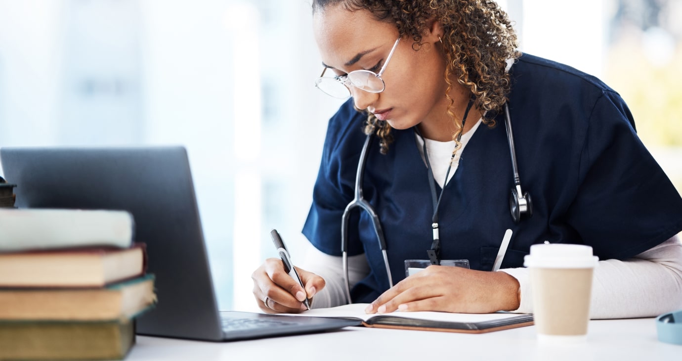 A person in medical scrubs writes in a notebook at a desk with a laptop, books, and a coffee cup, delving into best practices to enhance collaborative education within healthcare models.