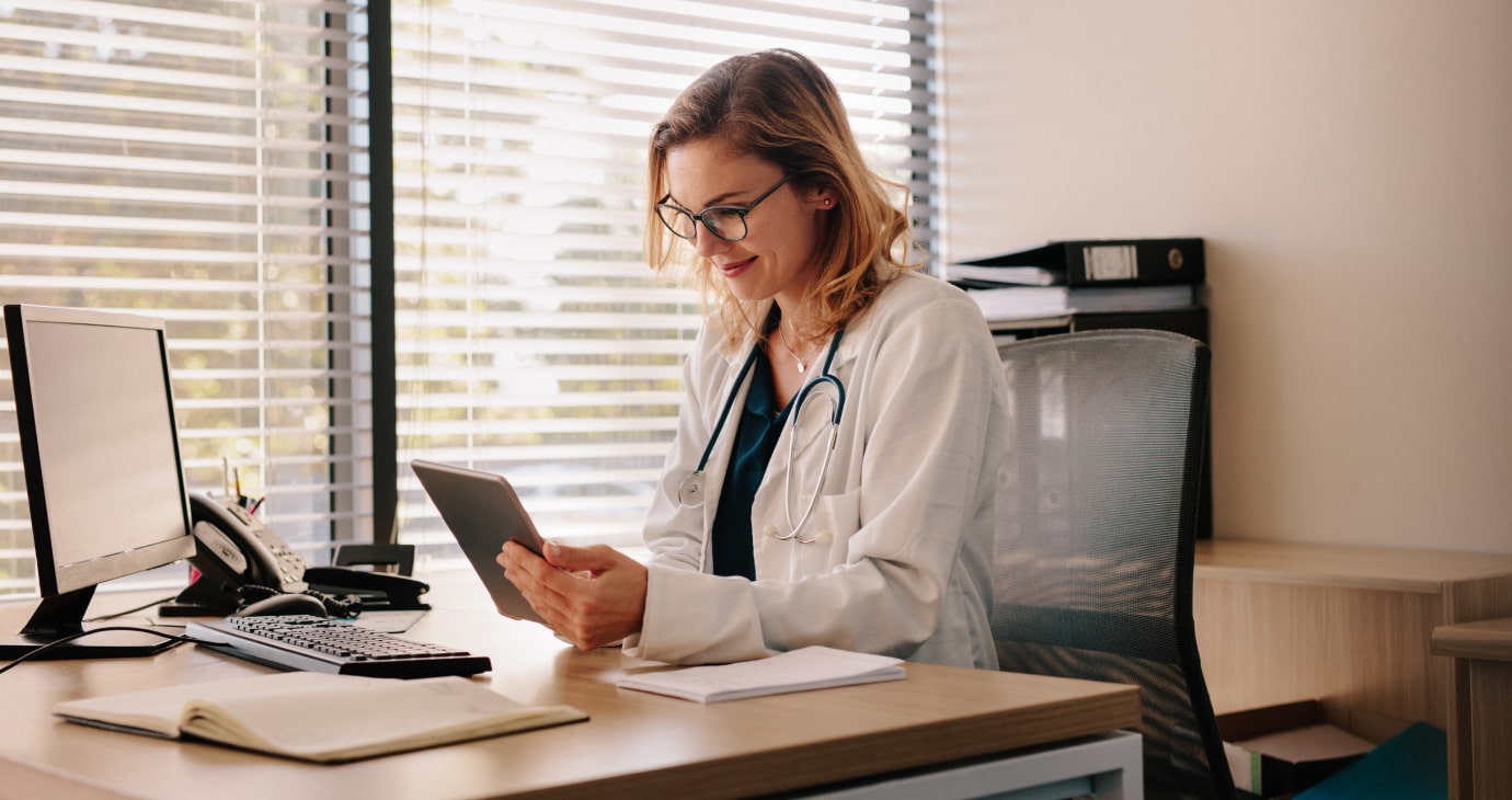 A healthcare professional in a white coat and stethoscope sits at a desk, focused on a tablet. A computer monitor, keyboard, and office items clutter the space as blinds cover the window in the background, illustrating the integration of modern education models in medical practice.