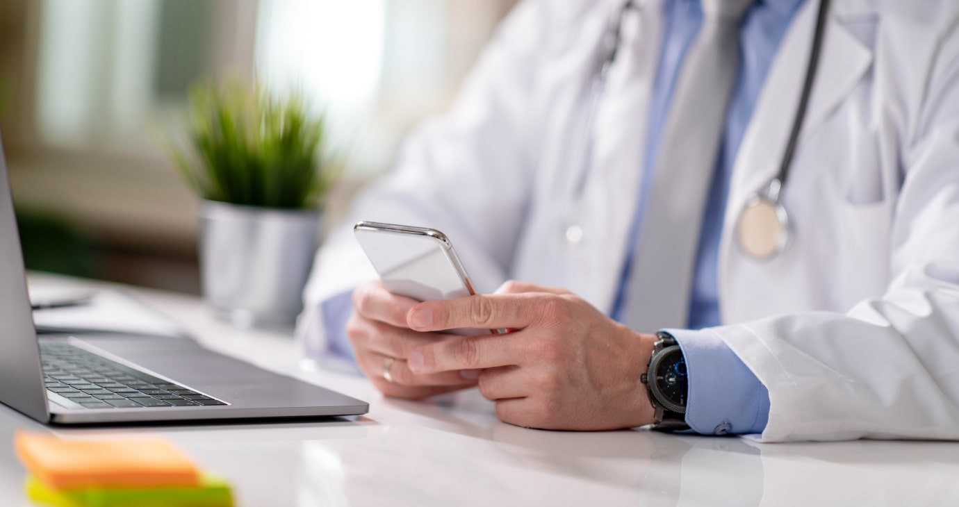 A physician in a white coat sits at a desk, smartphone in hand. Nearby, a laptop is open and a small potted plant adds a touch of nature, hinting at the balance between medical dedication and financial planning.