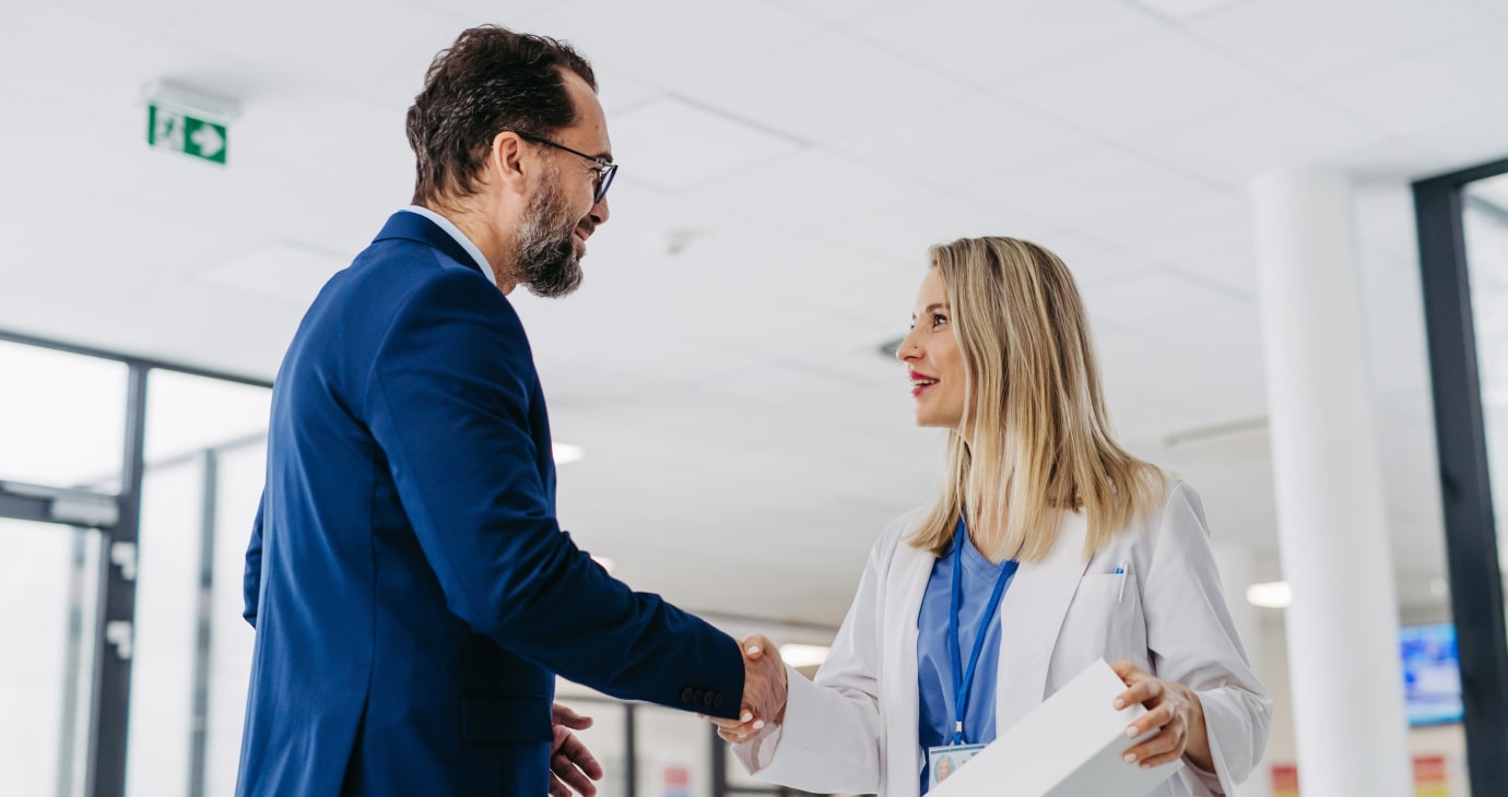 A man in a blue suit and a woman in a white coat with a lanyard, likely physicians, shake hands in a modern office setting.