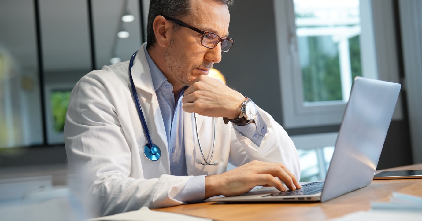 A physician in a white coat and stethoscope is seated at a desk, concentrating intently while using a laptop.