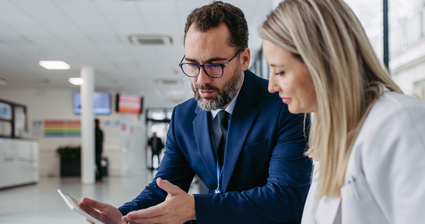 A man in a blue suit and a woman in a white coat discuss public health initiatives over a tablet in a modern office space.