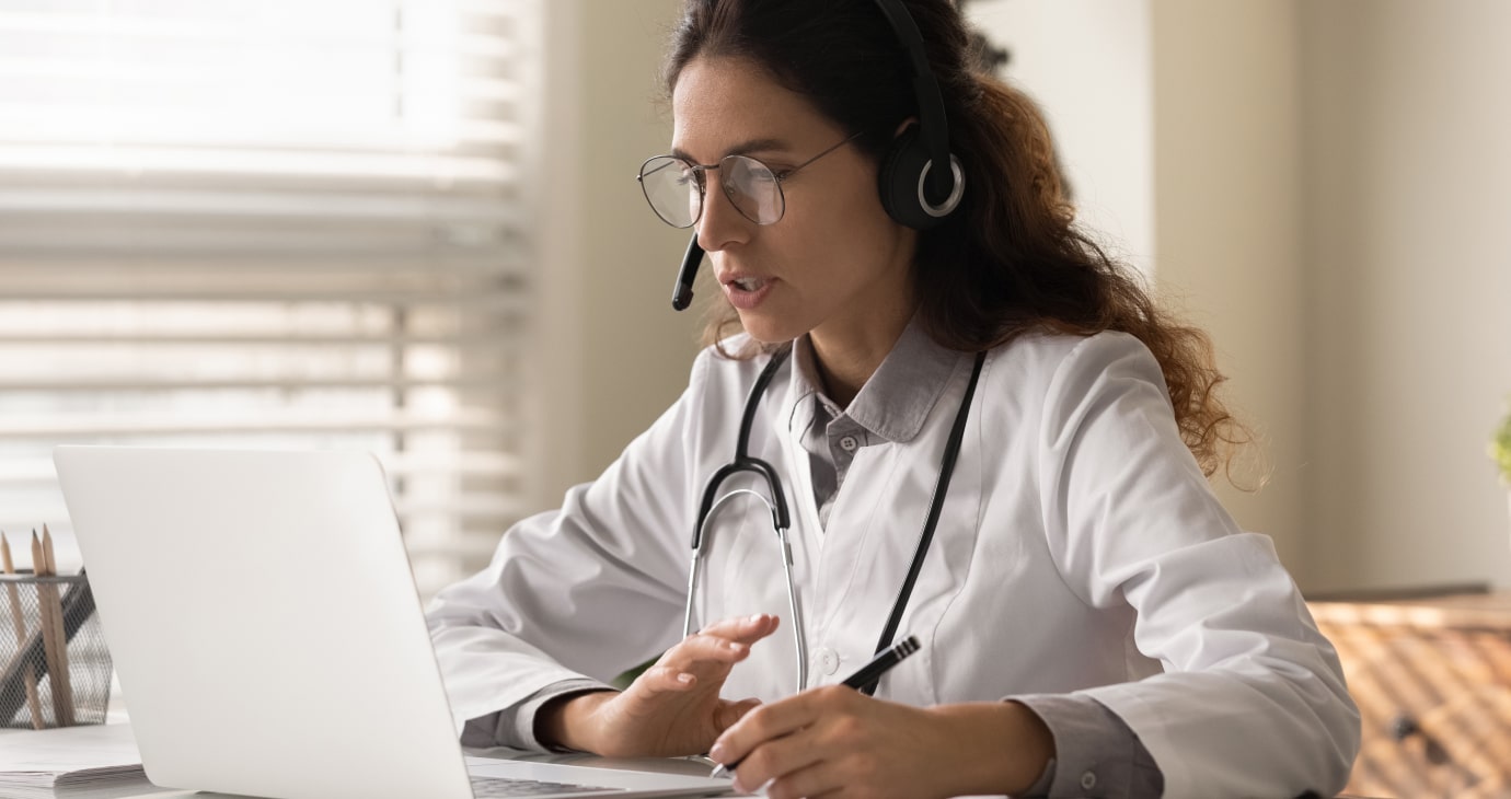 A woman in a white coat and stethoscope, embodying the dedication of doctors, is using a laptop, wearing a headset, and speaking during a virtual consultation.