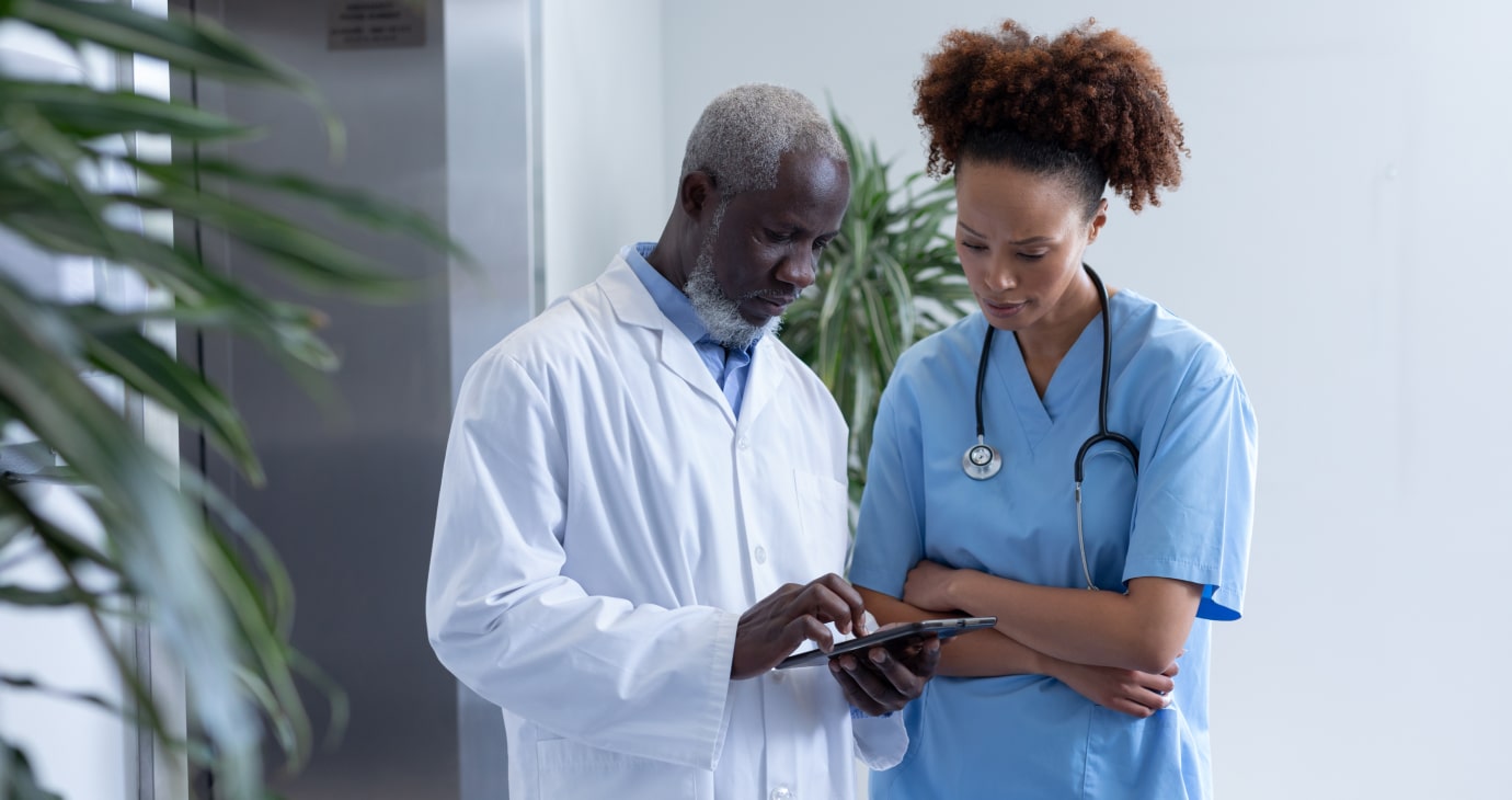 A man in a white lab coat, likely one of the hospital's doctors, shows a tablet to a woman in blue scrubs. They stand together in a hallway with plants, discussing crucial public health information.