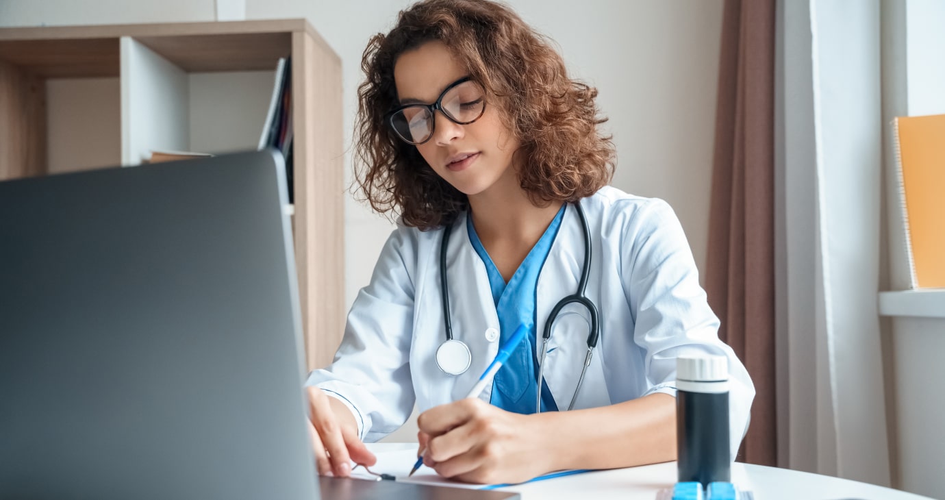 A doctor in a white coat and glasses is sitting at a desk, writing notes on paper while reviewing public health data on her laptop. A stethoscope hangs around her neck, a testament to policy research she frequently conducts, and a cup sits nearby.