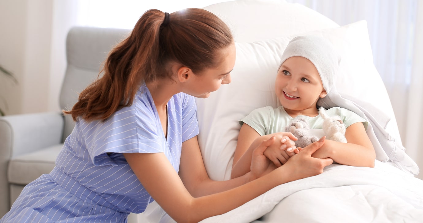 A woman in a blue striped dress sits beside a young girl wearing a headscarf in a hospital bed, holding her hand and smiling. They share hope as they discuss neoadjuvant therapies with their physicians, united in their fight against breast cancer.