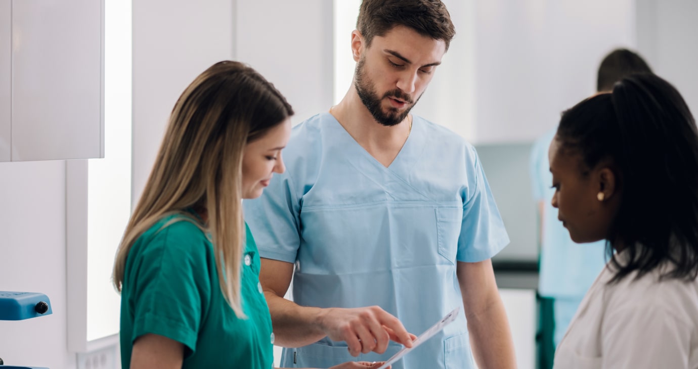 Three healthcare physicians are in discussion, one pointing at a document. They are wearing scrubs and standing in a clinical setting, likely reviewing neoadjuvant therapies related to breast cancer treatment options.