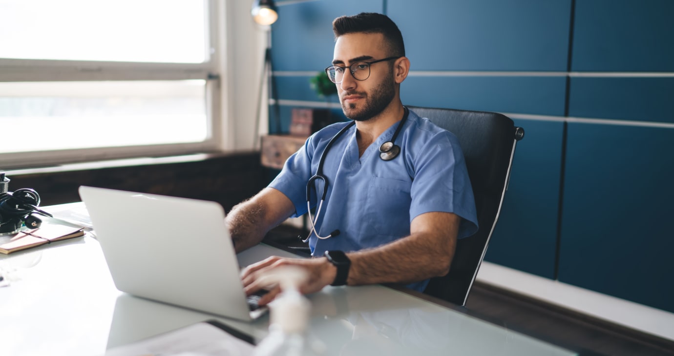 A physician wearing scrubs and a stethoscope diligently reviews Neoadjuvant therapies for breast cancer on his laptop in a well-lit office with a large window and blue wall.