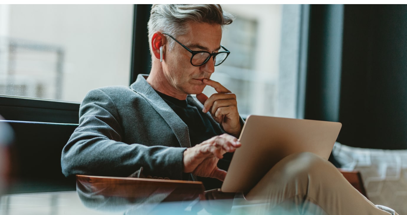 A man in glasses sits by the window, wearing a blazer and earphones, deeply engrossed in his laptop as he researches savings strategies.