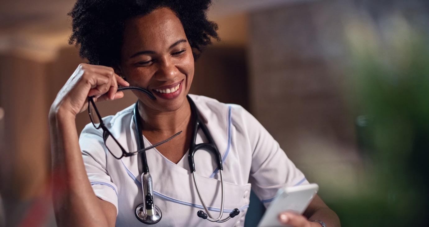 A smiling healthcare worker, focused on retirement advice, holds glasses while looking at a smartphone. With a stethoscope around the neck and wearing a white uniform, they balance daily tasks with long-term savings strategies for the future.