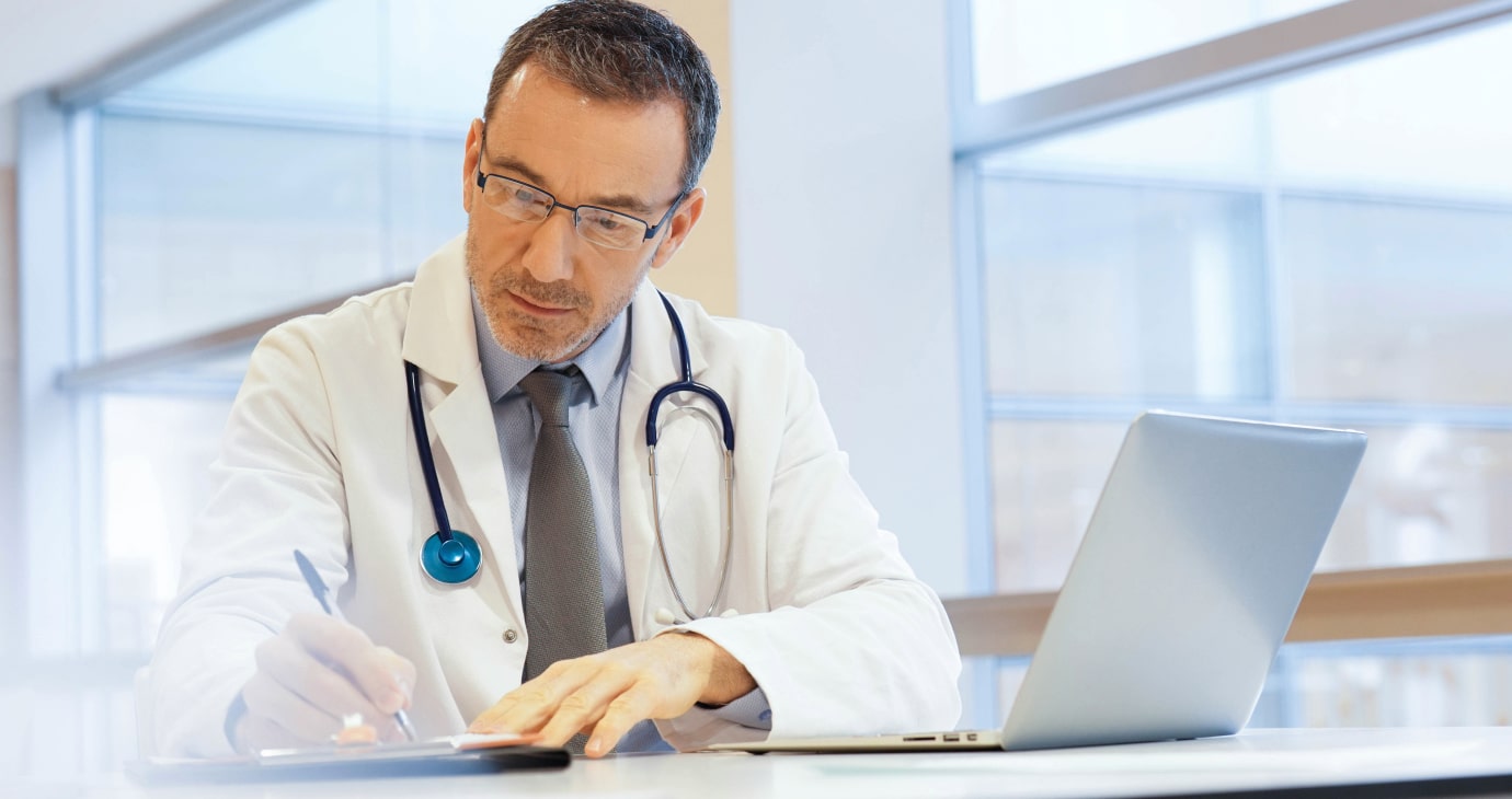 A doctor in a white coat with a stethoscope writes on a clipboard, focusing on retirement advice, while seated at a desk with a laptop in a bright office.