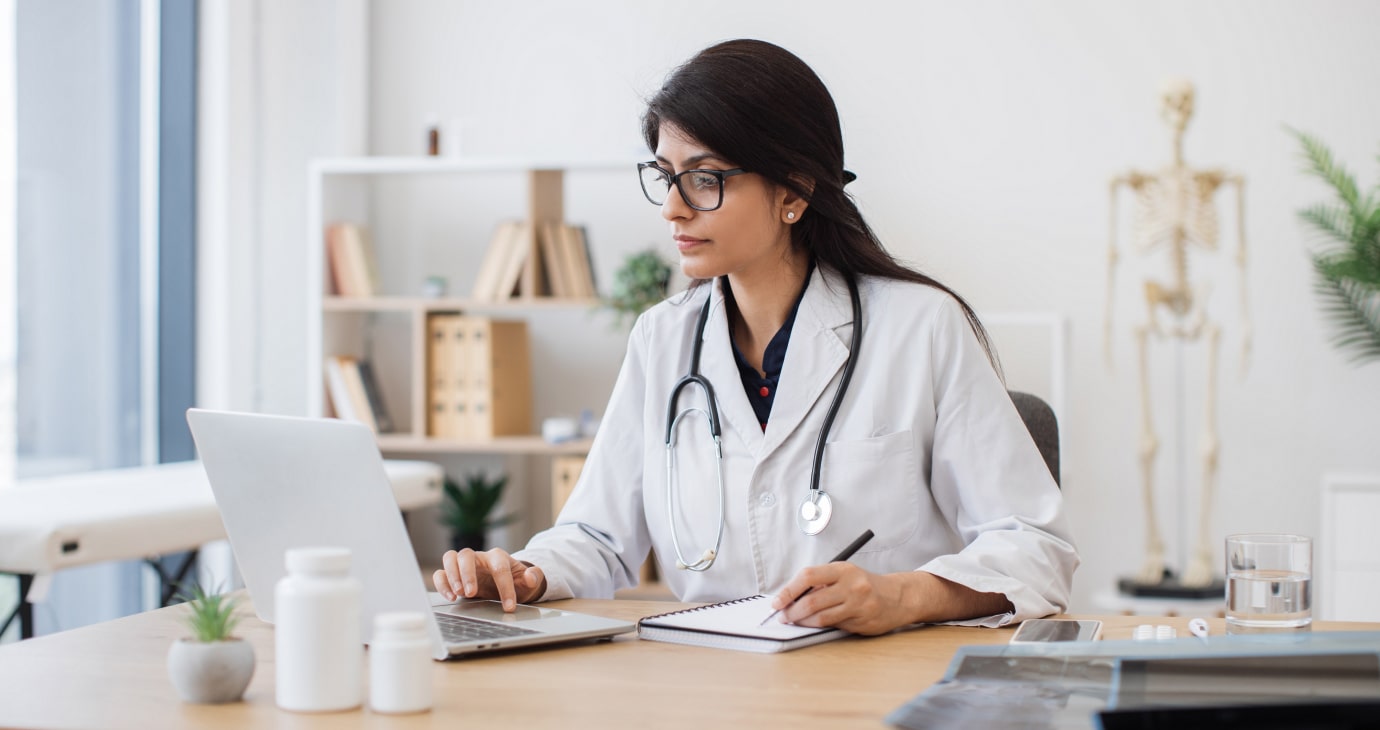 A doctor in a white coat and stethoscope sits at a desk, using a laptop to peruse a practical guide on physician contract review. As they jot down notes in their notebook, medical books and a skeleton model provide an academic backdrop.