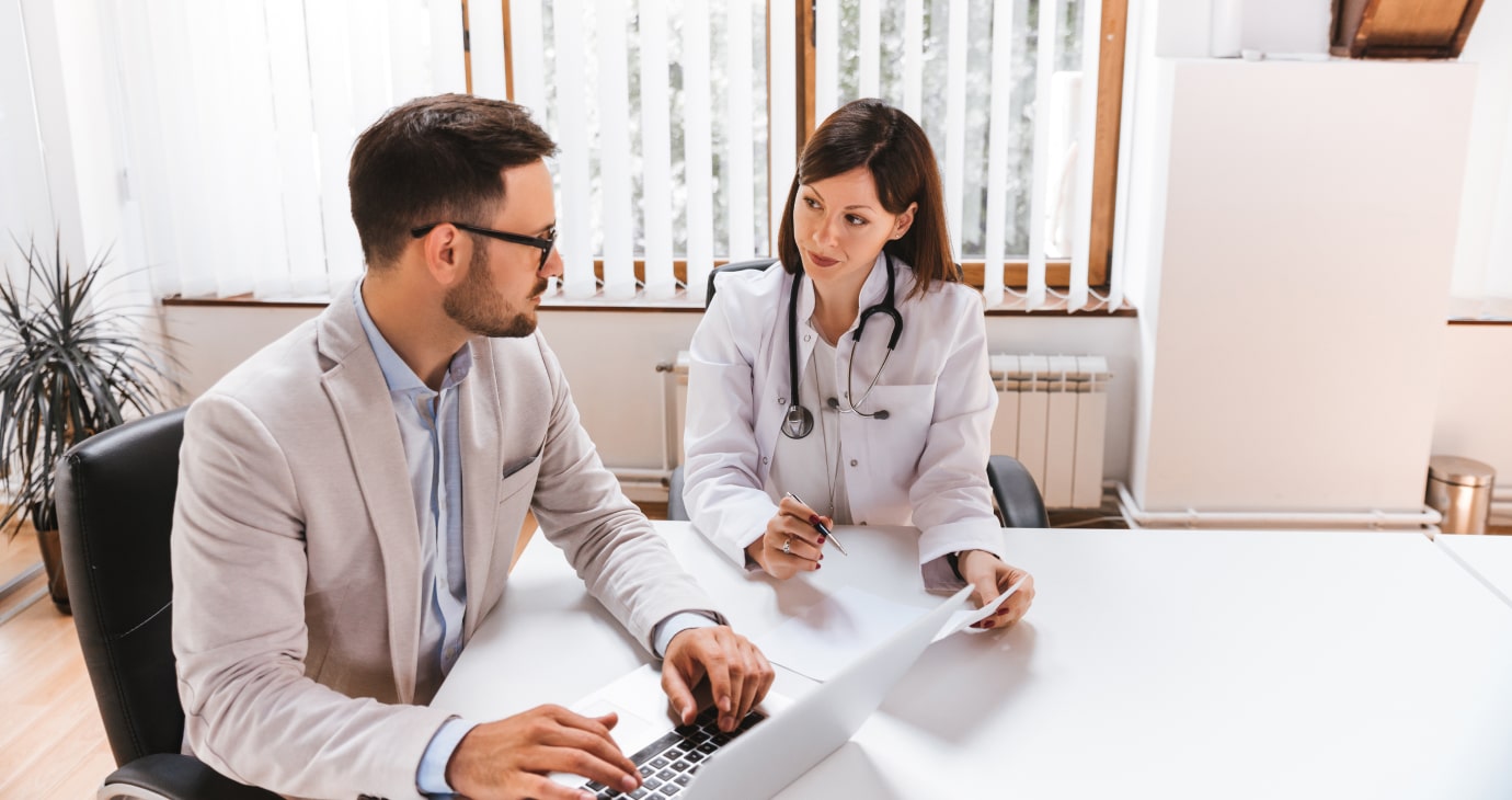 A doctor and a man in a suit sit at a desk in an office, engaged in a physician contract review. The doctor holds a pen and paper while the man diligently types on his laptop, consulting their practical guide for insights.