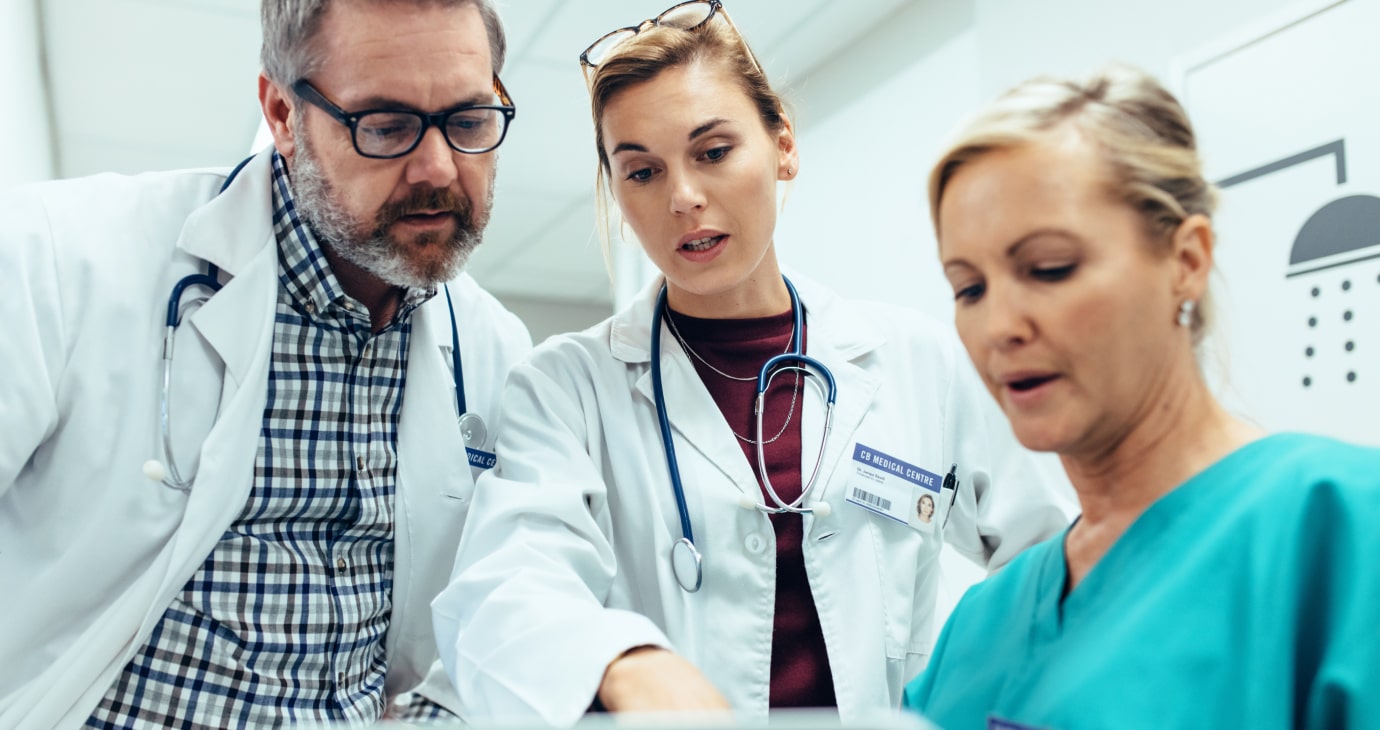 Three medical professionals review documents diligently; two in white coats with stethoscopes, one in scrubs. Their focus on physician contract details highlights their commitment to understanding this practical guide for better healthcare delivery.