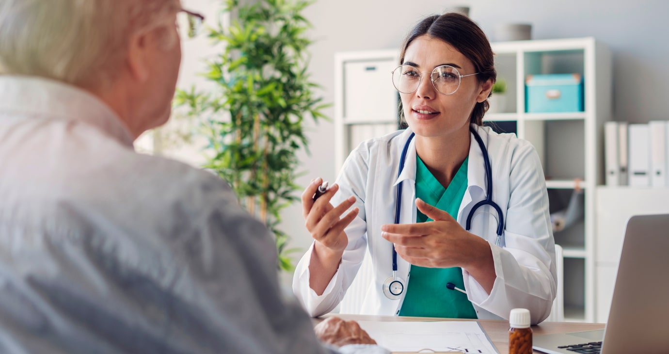 A physician in a white coat and stethoscope discusses healthcare options, including the rising relevance of Ozempic, with an older man across the table in a medical office setting.