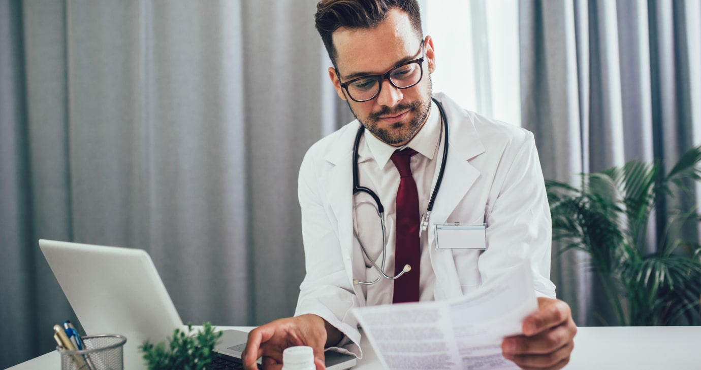 A physician in a white coat reviews a document at his desk, with a laptop open and a stethoscope draped around his neck. As interest in Ozempic continues to rise, he stays informed amid the serene backdrop of a green plant.