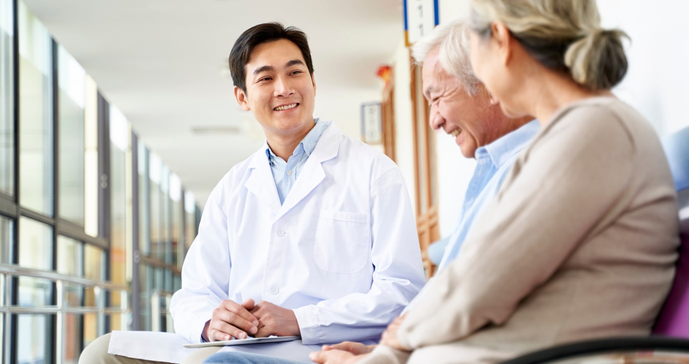 A smiling physician in a white coat talks to an elderly man and woman about pain management, creating a warm and friendly atmosphere in the hallway.