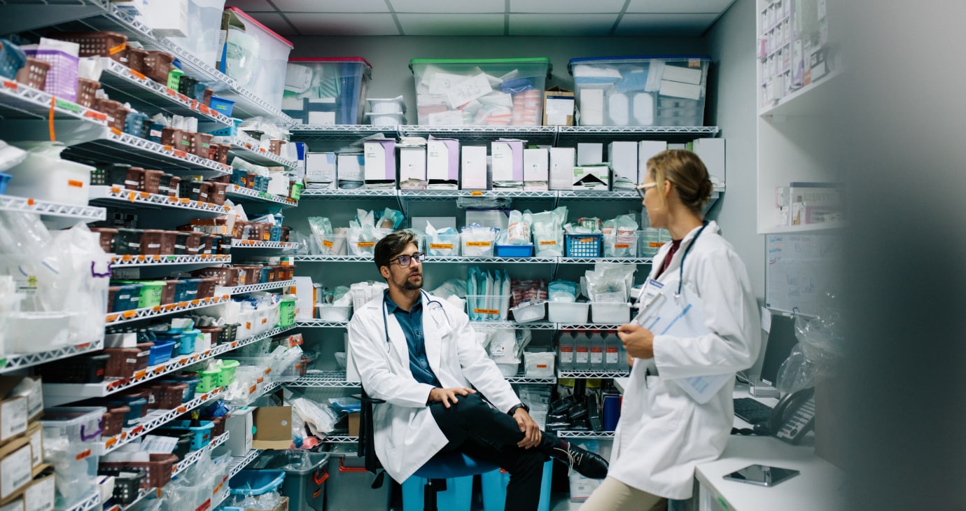 Two physicians in white coats converse in a well-stocked supply room, evaluating shelves brimming with various medical supplies and equipment crucial for end-of-life pain management.